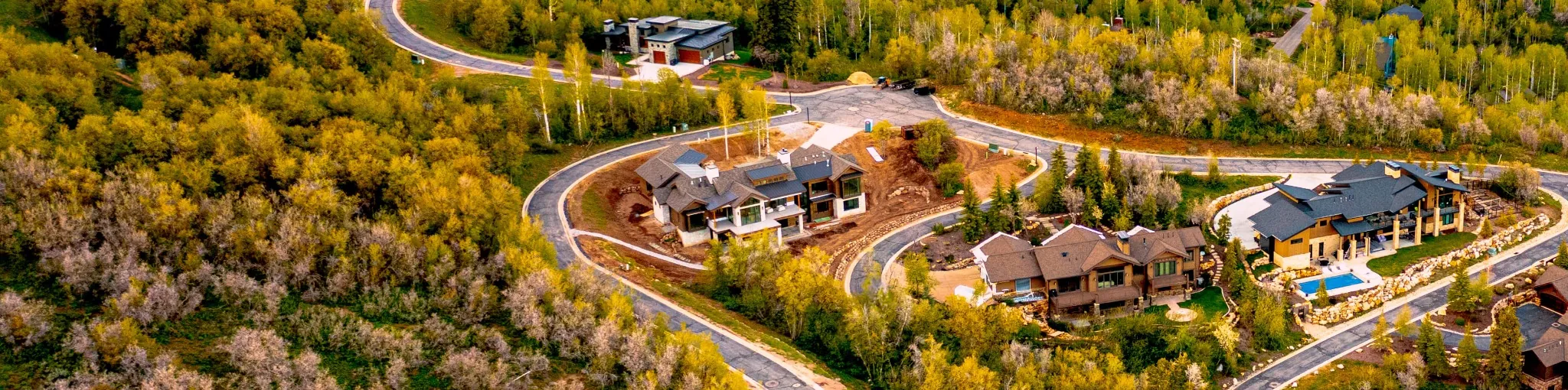Luxury custom homes under construction in a forested Park City neighborhood, surrounded by fall colors and winding mountain roads.